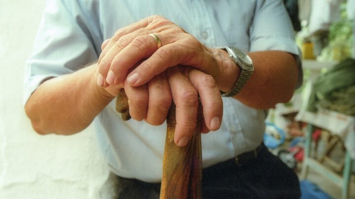 An older person sitting on a bench with a walking stick